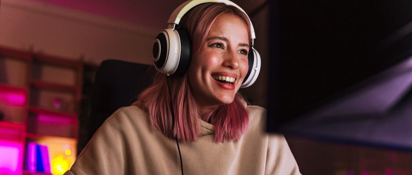 Image of joyful girl in headphones smiling and playing video game on computer while sitting at table indoors